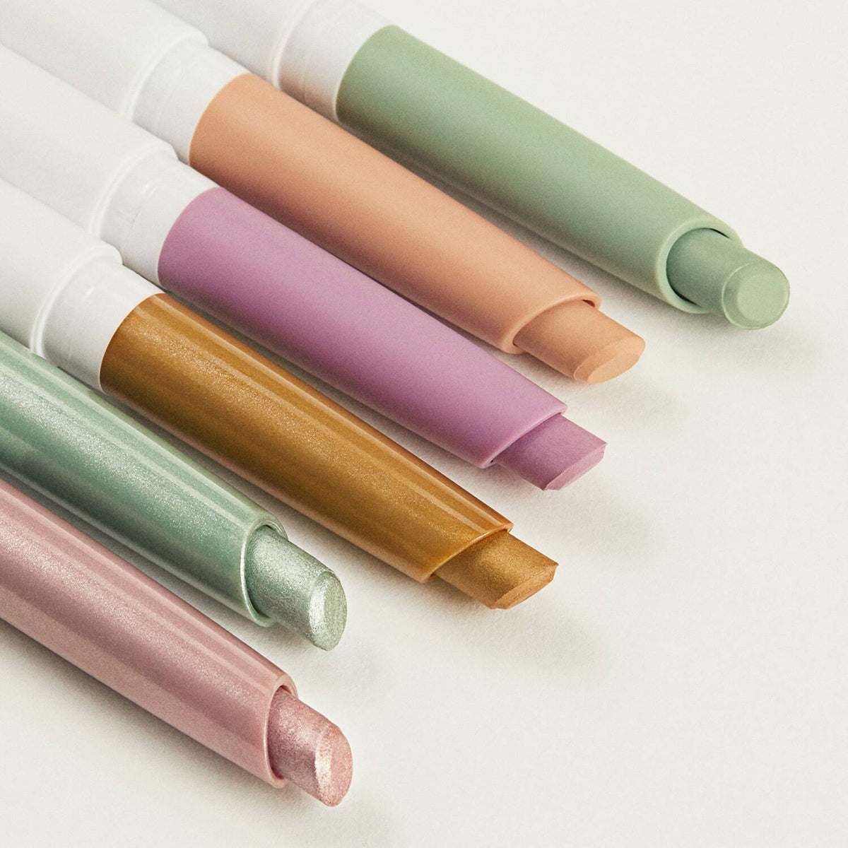 Four pastel lip glosses or lip balm tubes arranged diagonally on a clean white background, showing shades of sage green, peach, lavender, and metallic gold.