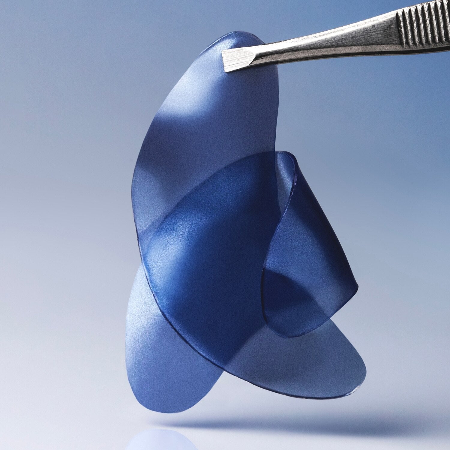 A translucent blue flexible film or sheet being held by metal tweezers against a soft blue gradient background.