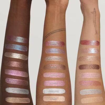 Three arms displaying vertical swatches of shimmering eyeshadow in various shades, including pinks, purples, silvers, and browns, against a light gray background.