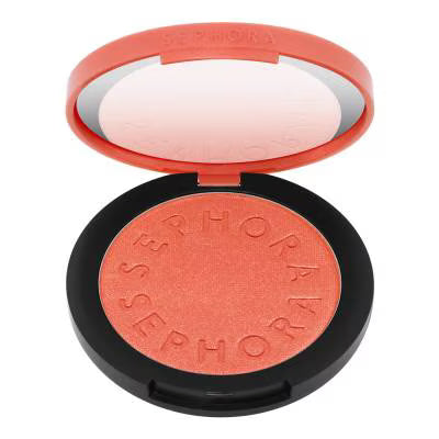 A round makeup compact with an open coral-red lid showing a mirror, and a pressed powder blush in a peachy coral shade inside a black case. The brand name 'SEPHORA' is embossed around the powder.