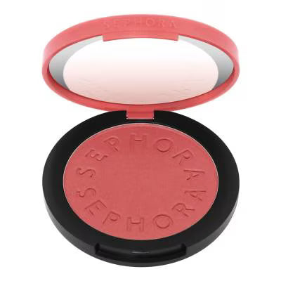 A round makeup compact with a black base and coral-pink lid, shown open to reveal a pressed blush in a matching coral-pink shade. The compact is embossed with the brand name 'SEPHORA'.