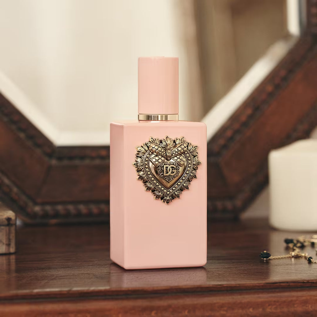 A pink rectangular perfume bottle with a matching pink cap and a decorative metallic heart emblem on the front, displayed on a wooden surface with a softly blurred background.
