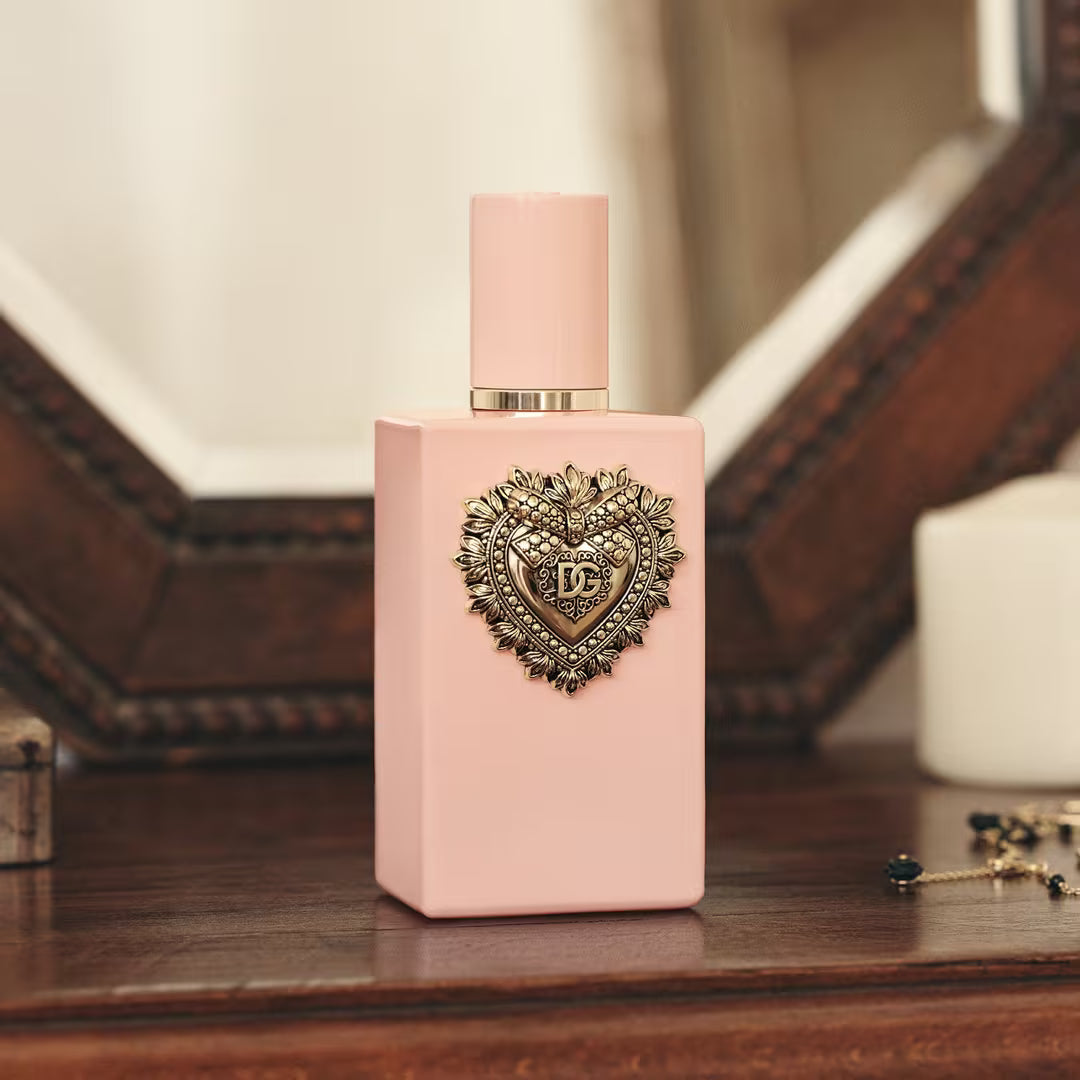 A pink rectangular perfume bottle with a matching pink cap and a decorative metallic heart emblem on the front, displayed on a wooden surface with a softly blurred background.
