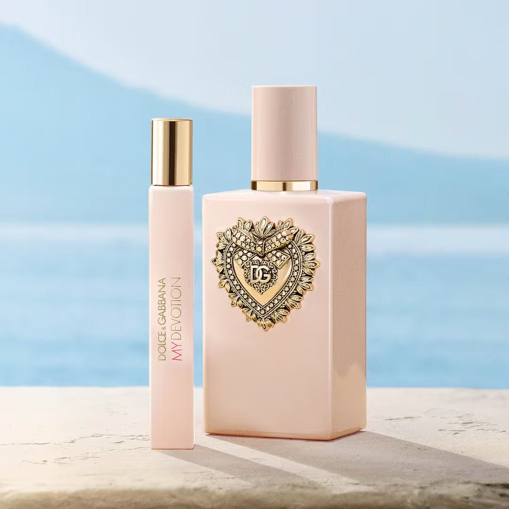 Dolce & Gabbana My Deoion perfume bottle and matching rollerball vial in a soft blush pink color with gold accents, set against a blurred blue ocean and mountain background.