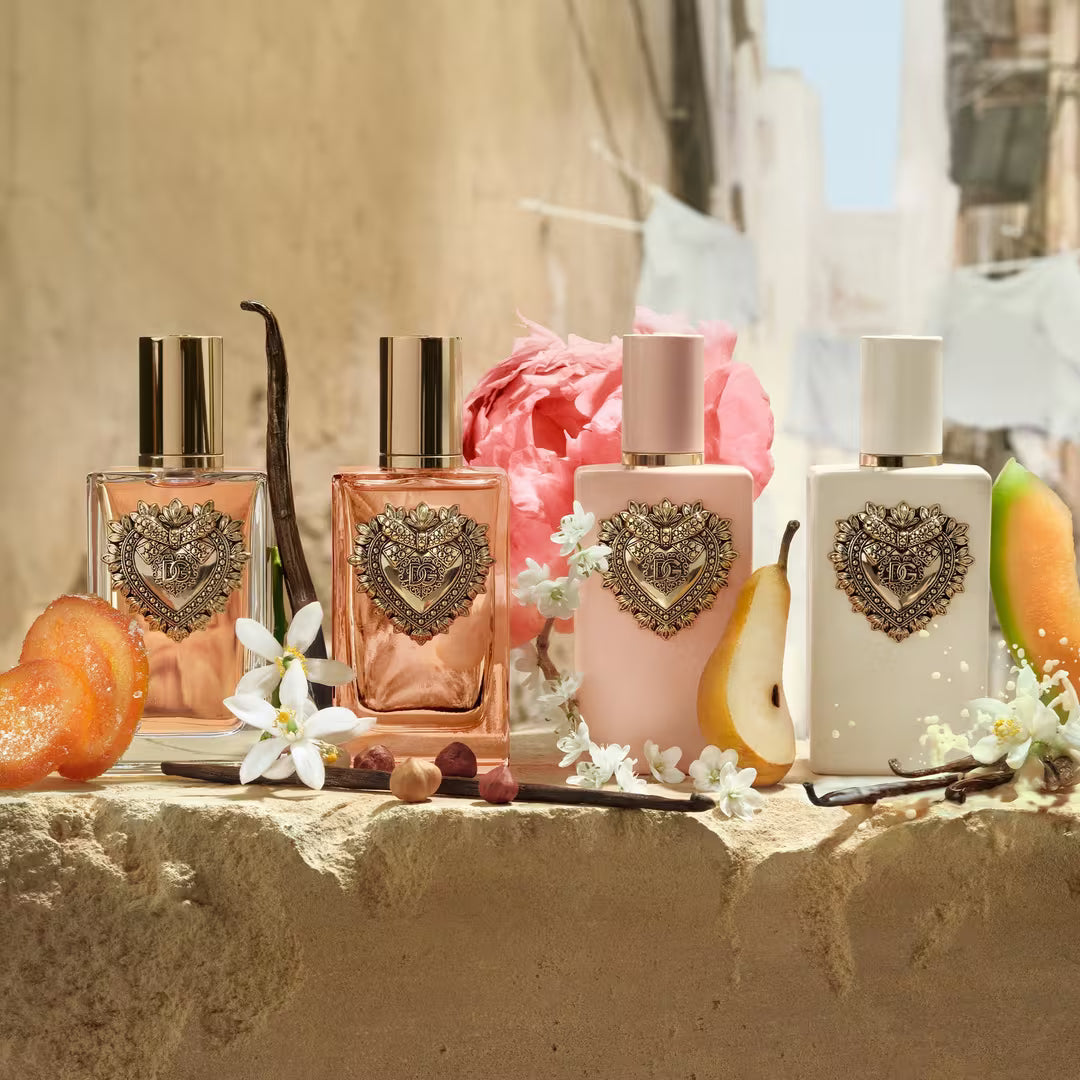 Four perfume bottles arranged on a stone surface outdoors, styled with flowers, fruit slices, and vanilla beans. The bottles are pink, peach, pale pink, and white with ornate heart-shaped metallic emblems on the front.