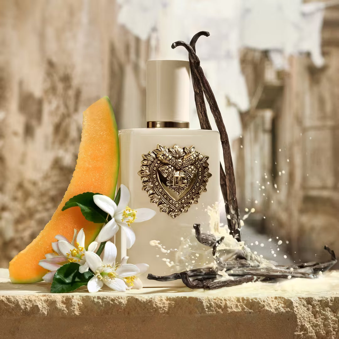 A perfume bottle with a decorative gold crest on a beige surface, styled with a cantaloupe melon slice, white flowers, vanilla beans, and a splash of liquid against a blurred rustic background.