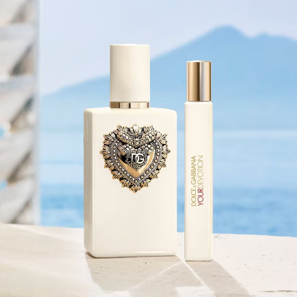 Dior Scent of Liberty perfume bottle in off-white with a gold and crystal heart emblem, shown next to a matching travel atomizer spray bottle against a blurred blue ocean and mountain background.