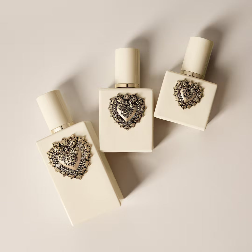 Three cream-colored square cosmetic bottles with ornate heart-shaped silver metal embellishments on the front, arranged on a light beige background.