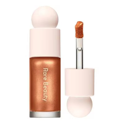 Rare Beauty liquid highlighter in a metallic copper-rose shade, shown with the cap off and the applicator wand beside it on a white background.