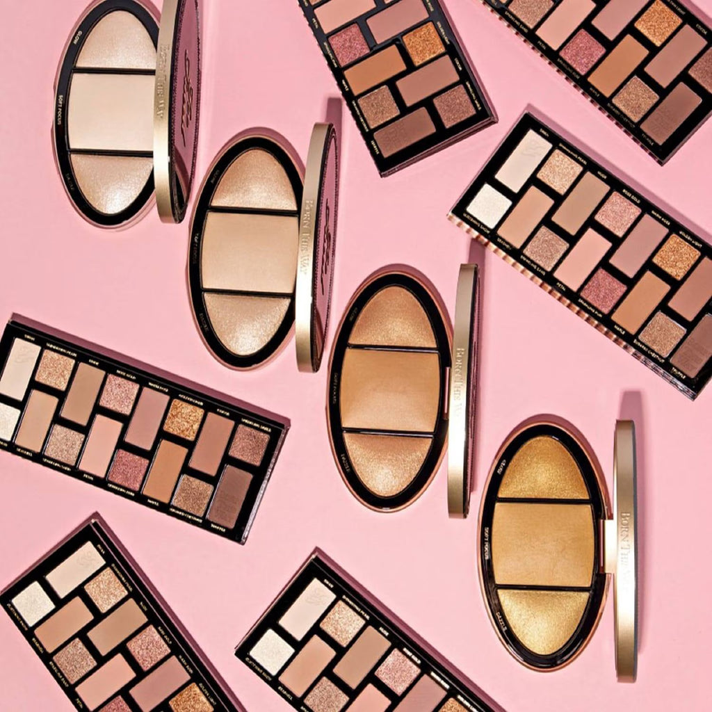 An overhead flat-lay arrangement of multiple makeup palettes and compacts on a solid pink background. The products include rectangular and oval eyeshadow palettes with neutral, bronze, and gold shades, alongside shimmer highlighters and cream products.