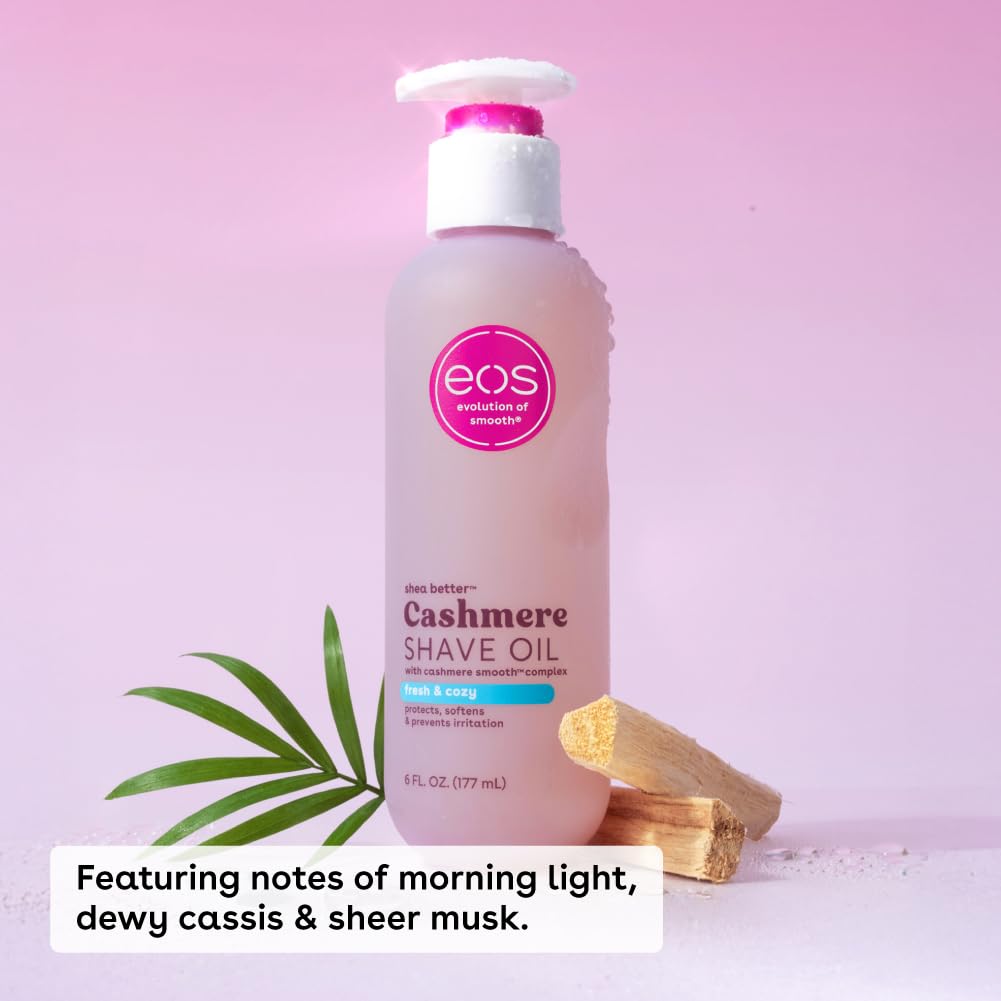A bottle of EOS Cashmere Shave Oil with a white pump and pink cap, set against a soft pink background with decorative green leaves and wooden wax melts. A text overlay at the bottom reads: 'Featuring notes of morning light, dewy cassis & sheer musk.