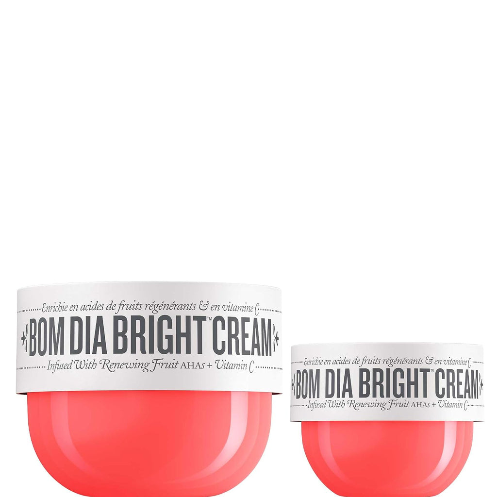 Two jars of BOM DIA BRIGHT CREAM on a plain white background, one larger jar in the foreground and one smaller jar behind it. The containers are translucent coral-pink with white labels featuring black text.