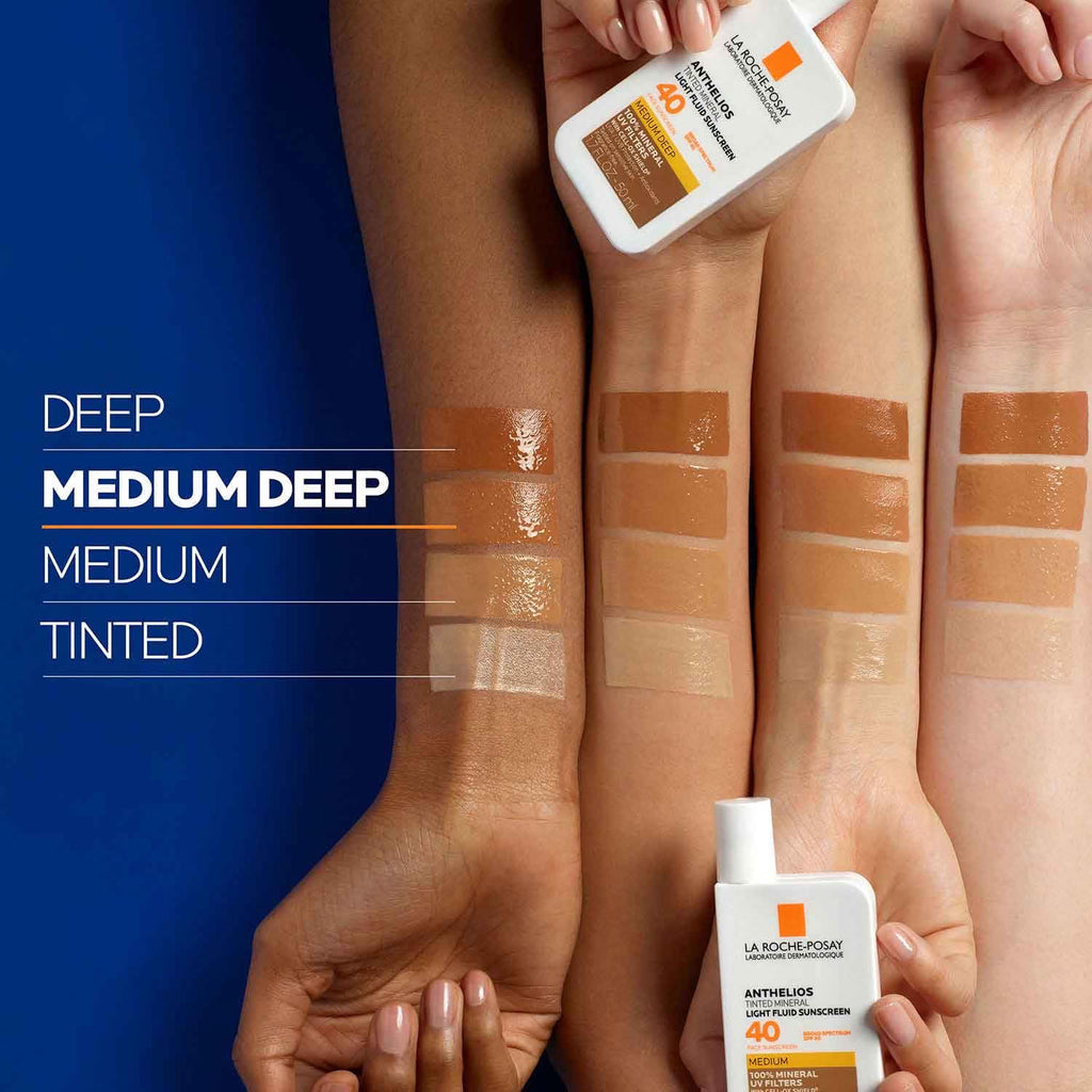 Hands holding bottles of sunscreen with color swatches on skin against a blue background