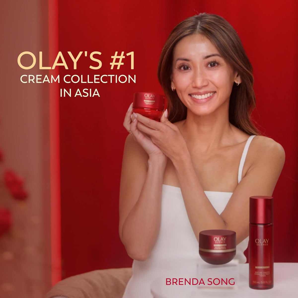 A smiling woman in a white dress holds an Olay cream jar while standing in front of a red backdrop, with additional Olay products on a table and promotional text reading 'OLAY'S #1 CREAM COLLECTION IN ASIA' and 'BRENDA SONG'.