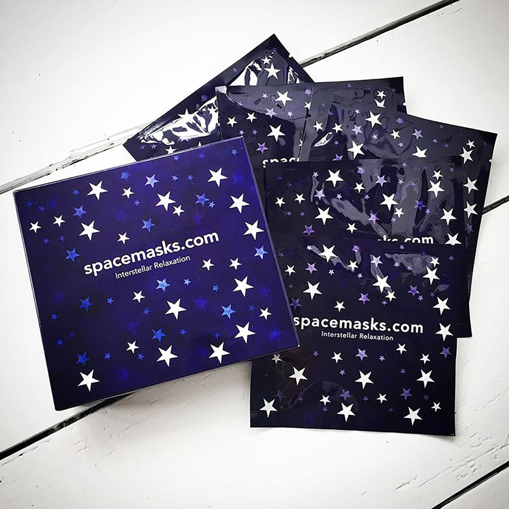 A boxed set of space-themed face masks from spacemasks.com, featuring a dark blue box and several individual packets decorated with white and light blue stars, arranged on a white wooden surface.