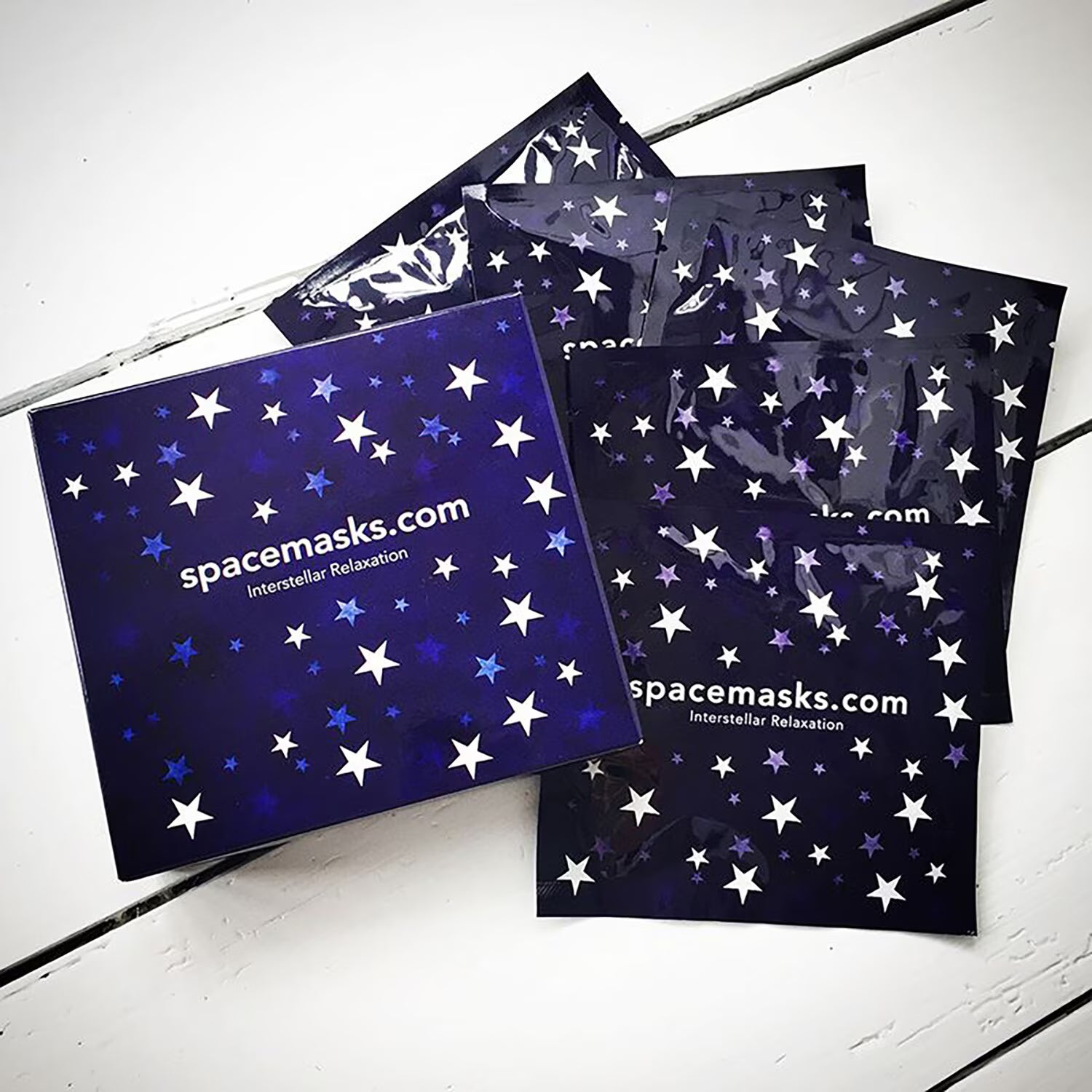A boxed set of space-themed face masks from spacemasks.com, featuring a dark blue box and several individual packets decorated with white and light blue stars, arranged on a white wooden surface.