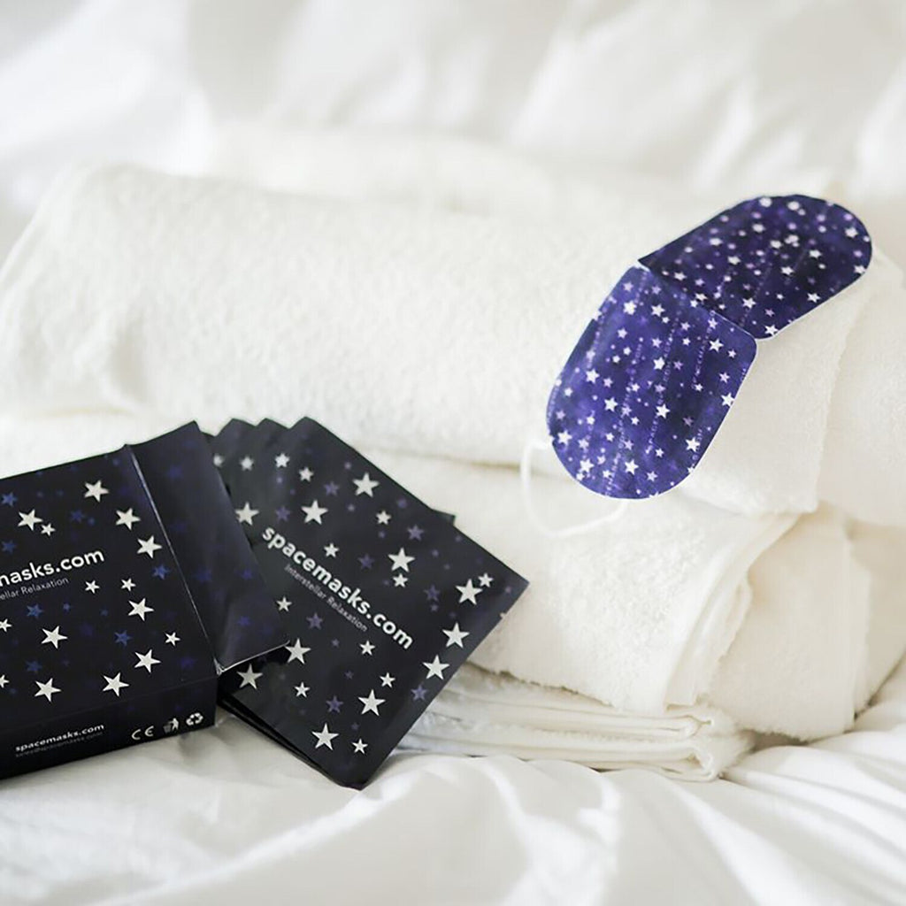 Two oval sleep masks with a navy blue and white star pattern are displayed on white towels next to matching star-patterned product packaging on a white bed.