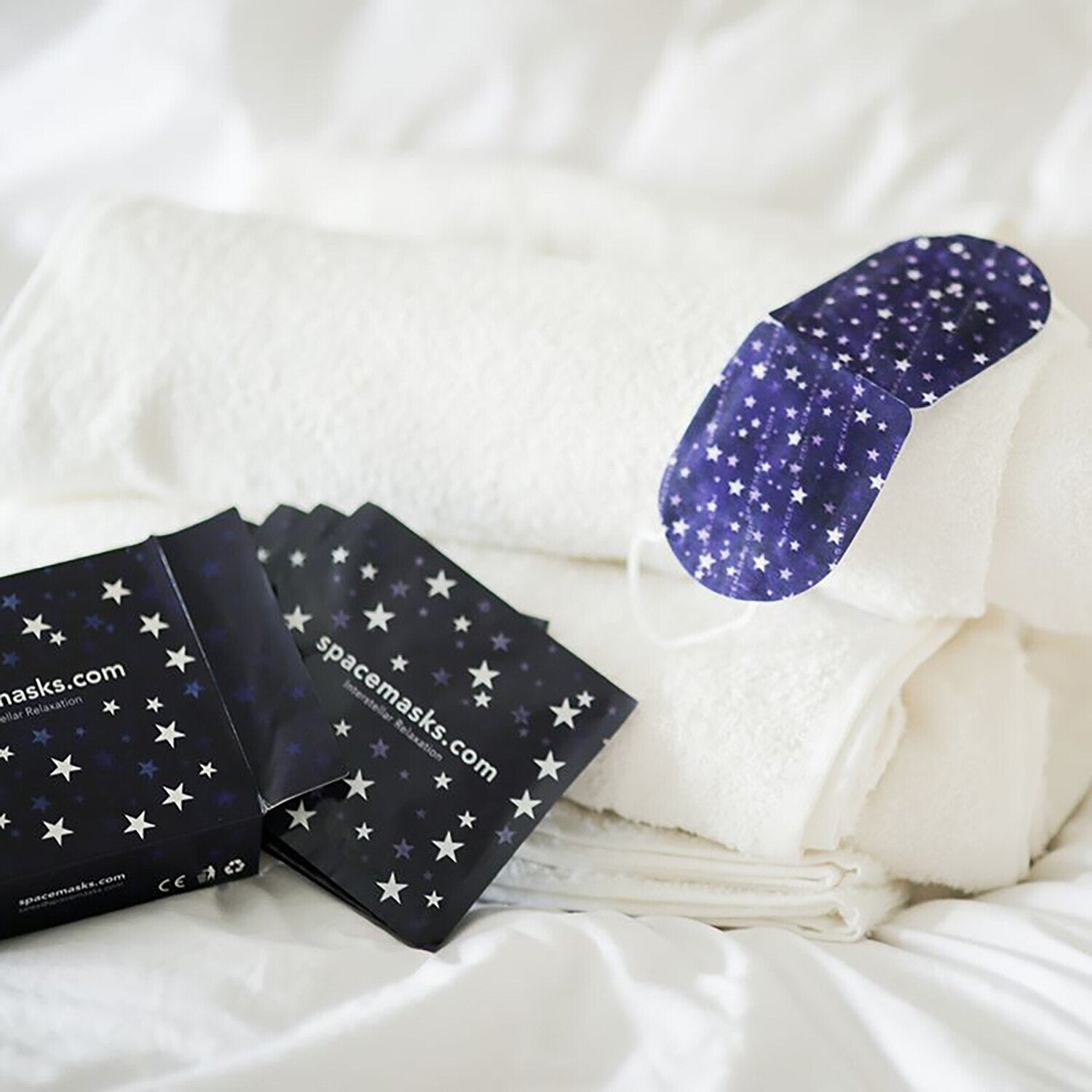 Two oval sleep masks with a navy blue and white star pattern are displayed on white towels next to matching star-patterned product packaging on a white bed.