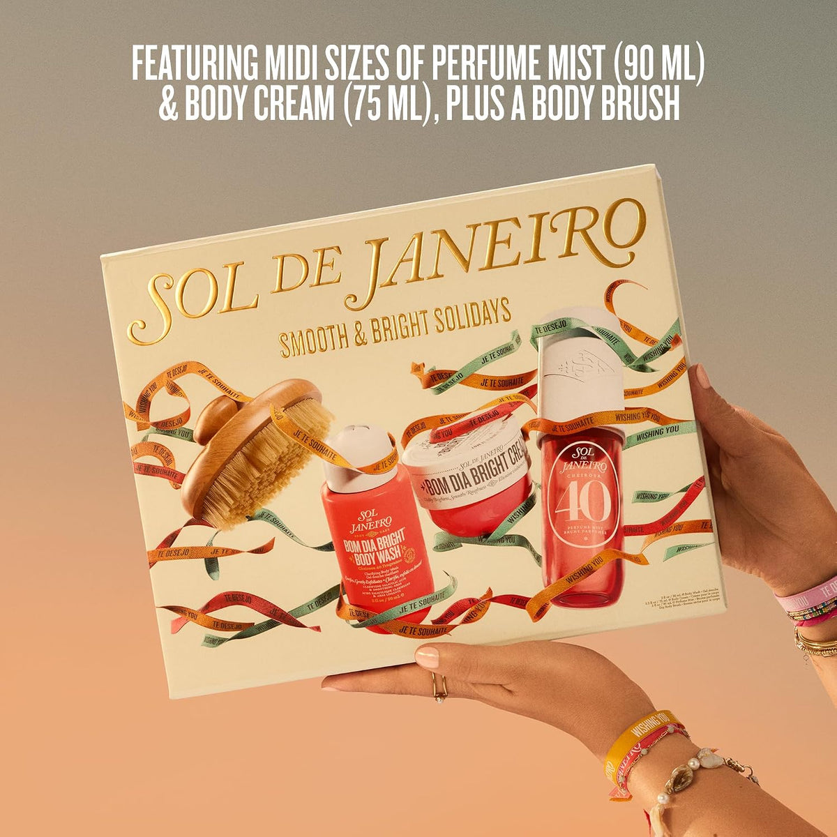 A gift set box held by two hands, featuring the brand name 'SOL DE JANEIRO' and holiday-themed illustrations of body care products and festive ribbons. Text at the top reads 'FEATURING MIDI SIZES OF PERFUME MIST (90 ML) & BODY CREAM (75 ML), PLUS A BODY BRUSH'.