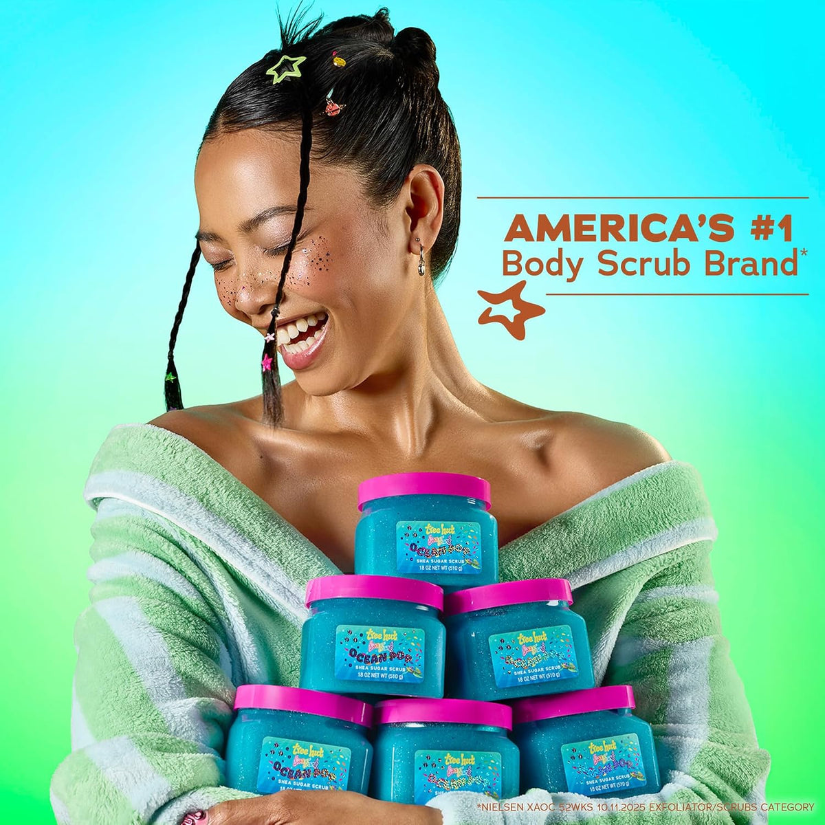 A woman wrapped in a light green towel smiles while holding a stack of blue body scrub jars with pink lids. The background is a bright green-to-blue gradient, and promotional text on the right reads 'AMERICA'S #1 Body Scrub Brand' with a star graphic.