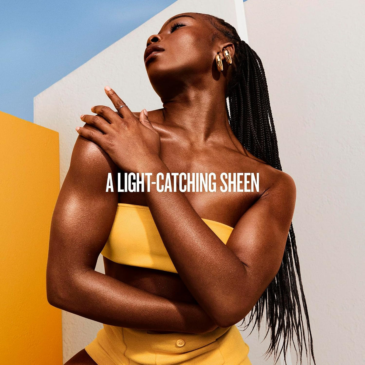 A woman with long black box braids poses outdoors against a geometric background of white and yellow walls under a blue sky. She is wearing a yellow strapless top and matching bottoms, with white text overlaid across the center reading 'A LIGHT-CATCHING SHEEN'.