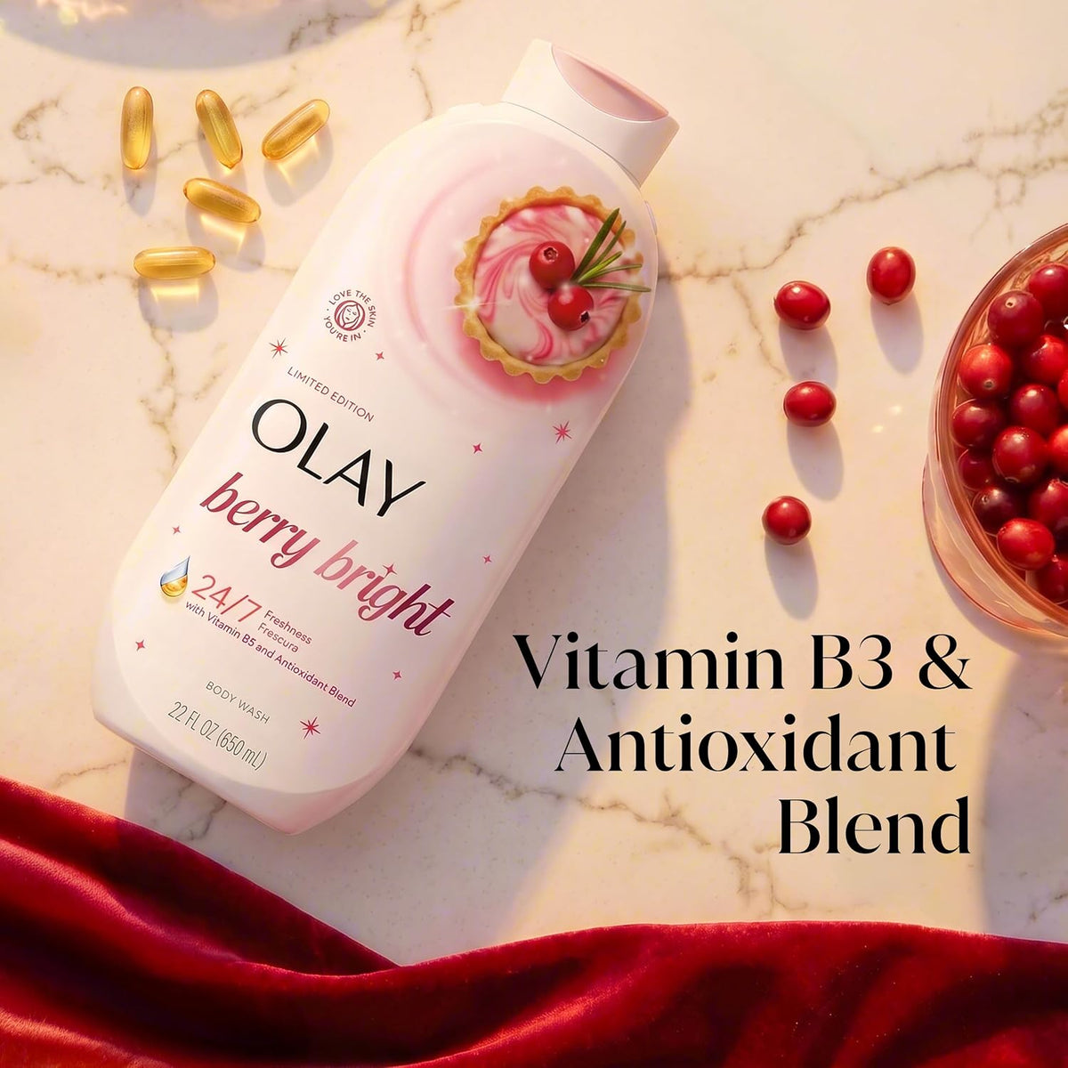 Olay Berry Bright Vitamin B3 & Antioxidant Blend body wash bottle on a white marble surface with scattered red cranberries, gold capsules, and a red velvet cloth. Text overlay reads 'Vitamin B3 & Antioxidant Blend'.
