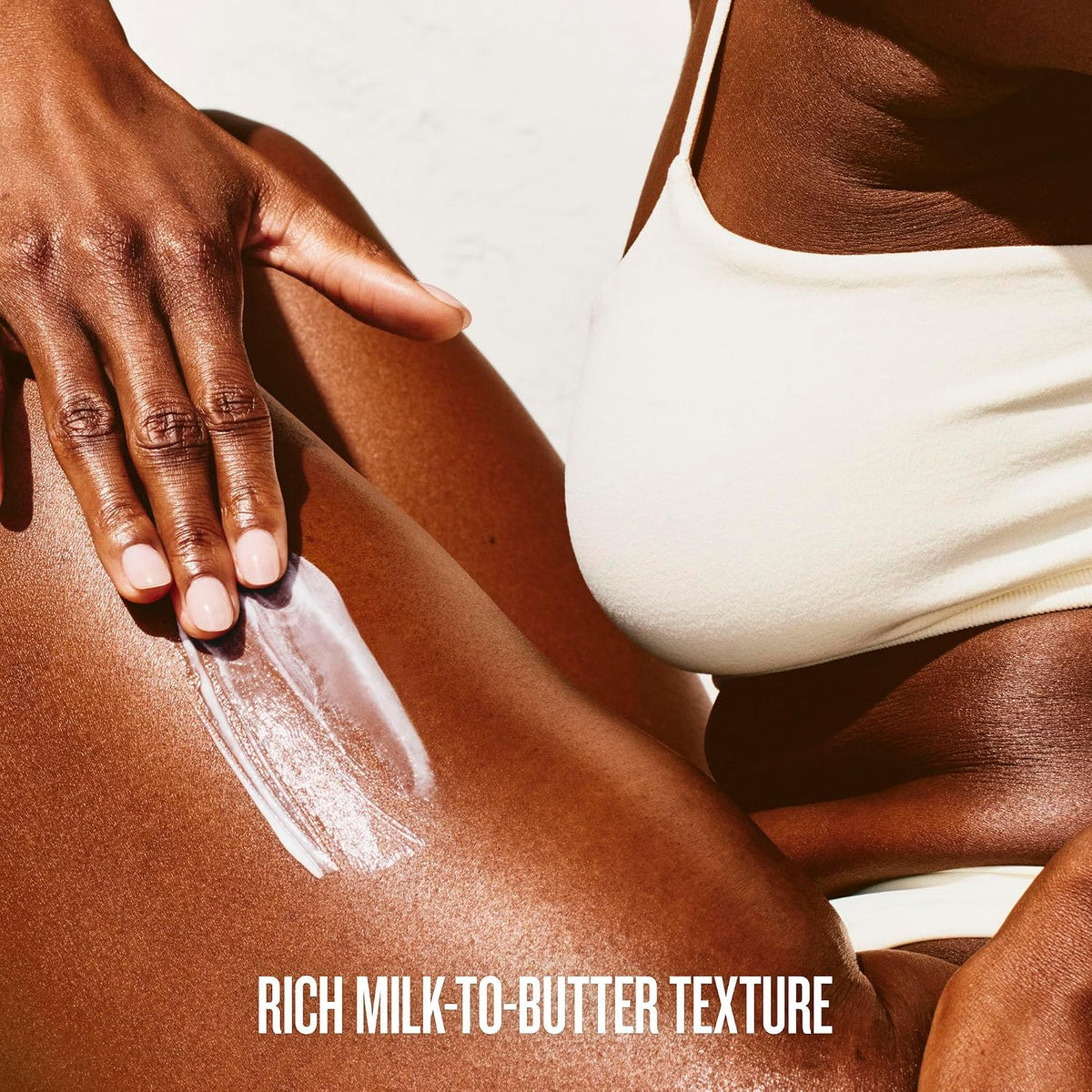 Close-up of a person applying a white cream or sunscreen lotion onto their leg, wearing a white bikini top. The text 'RICH MILK-TO-BUTTER TEXTURE' is displayed at the bottom.
