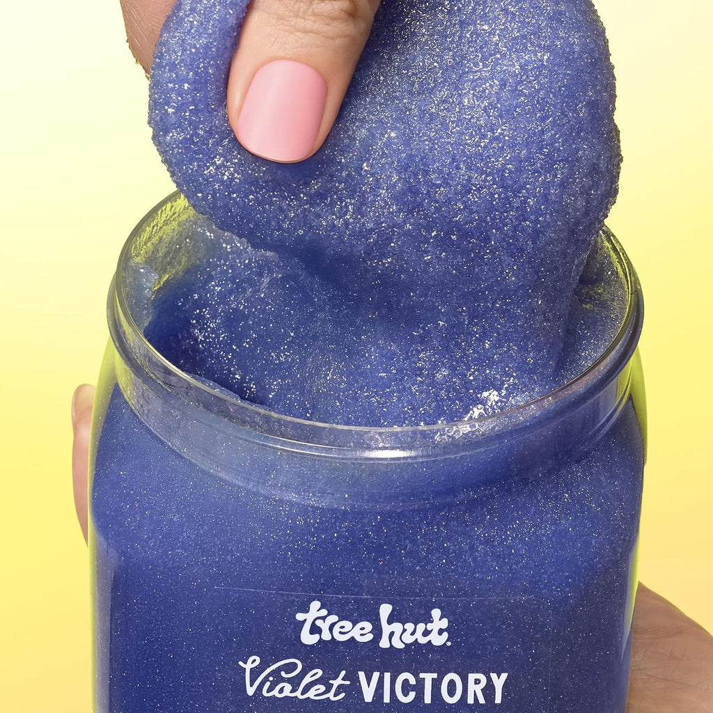 A close-up of a hand lifting a blue glittery, textured slime-like product out of a clear jar labeled 'tree hut Violet VICTORY' against a pale yellow background.