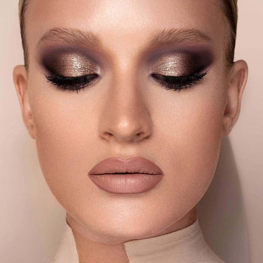 Close-up of a model with closed eyes wearing dramatic metallic eyeshadow in bronze, brown, and purple tones, with long black eyelashes and nude pink lipstick against a soft beige background.