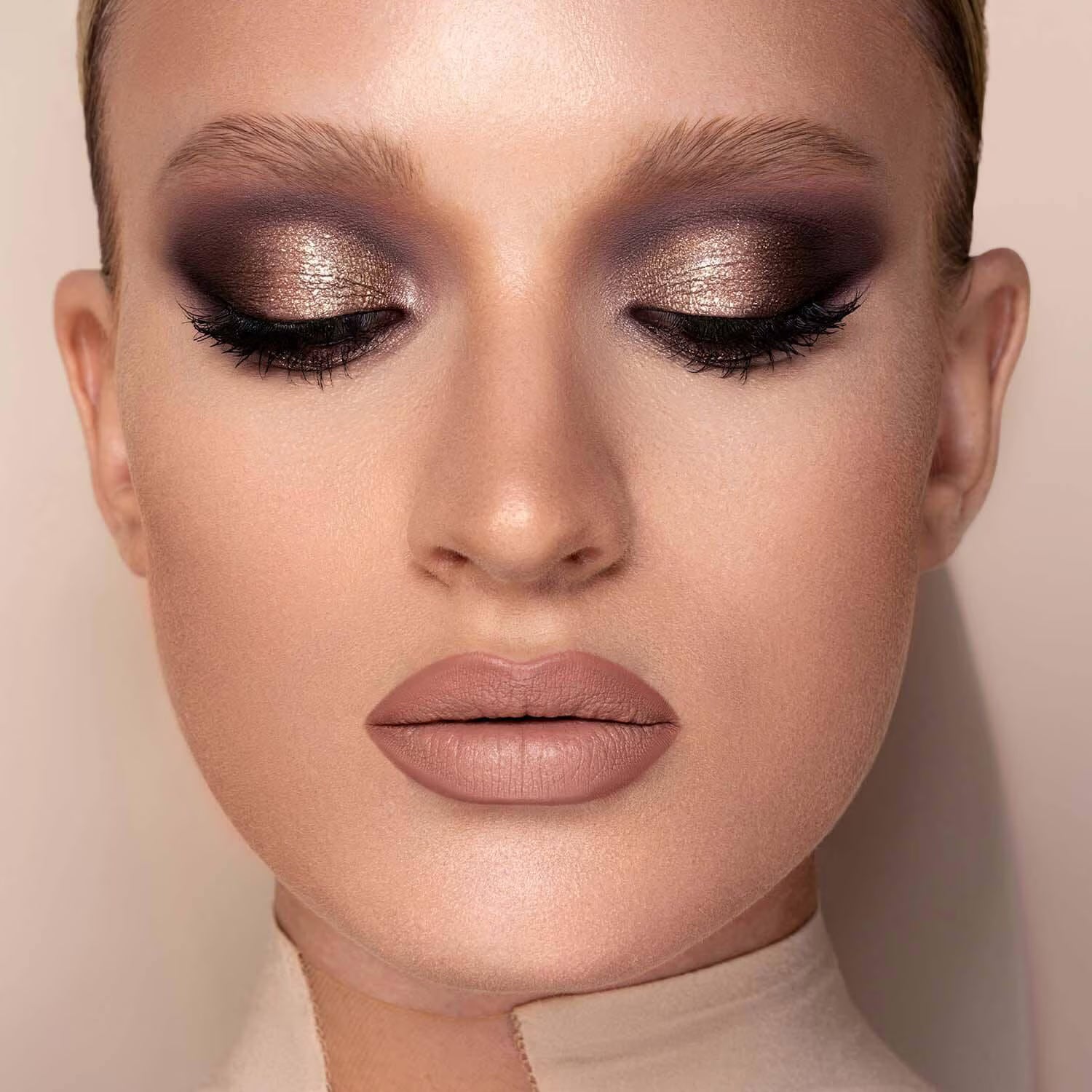 Close-up of a model with closed eyes wearing dramatic metallic eyeshadow in bronze, brown, and purple tones, with long black eyelashes and nude pink lipstick against a soft beige background.