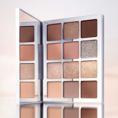 An open makeup palette with multiple square eyeshadow pans in warm neutral shades, including browns, tans, beiges, and a shimmery champagne tone, set against a soft light background.