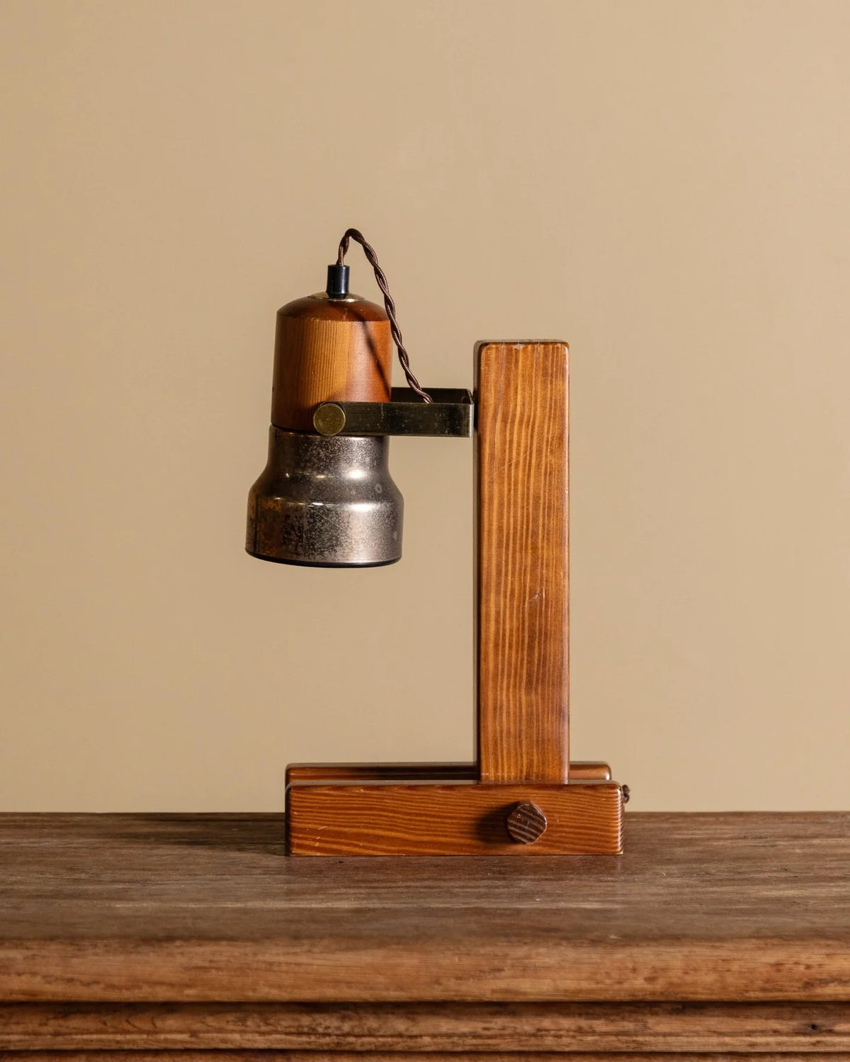 A rustic industrial-style table lamp with a weathered metal bell shade, a vertical wooden post, and a drawer base, set on a wooden surface against a beige background.