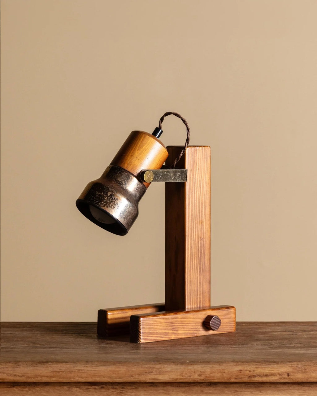 A rustic industrial-style table lamp with a wooden and metal body, shown on a wooden tabletop against a solid beige background.