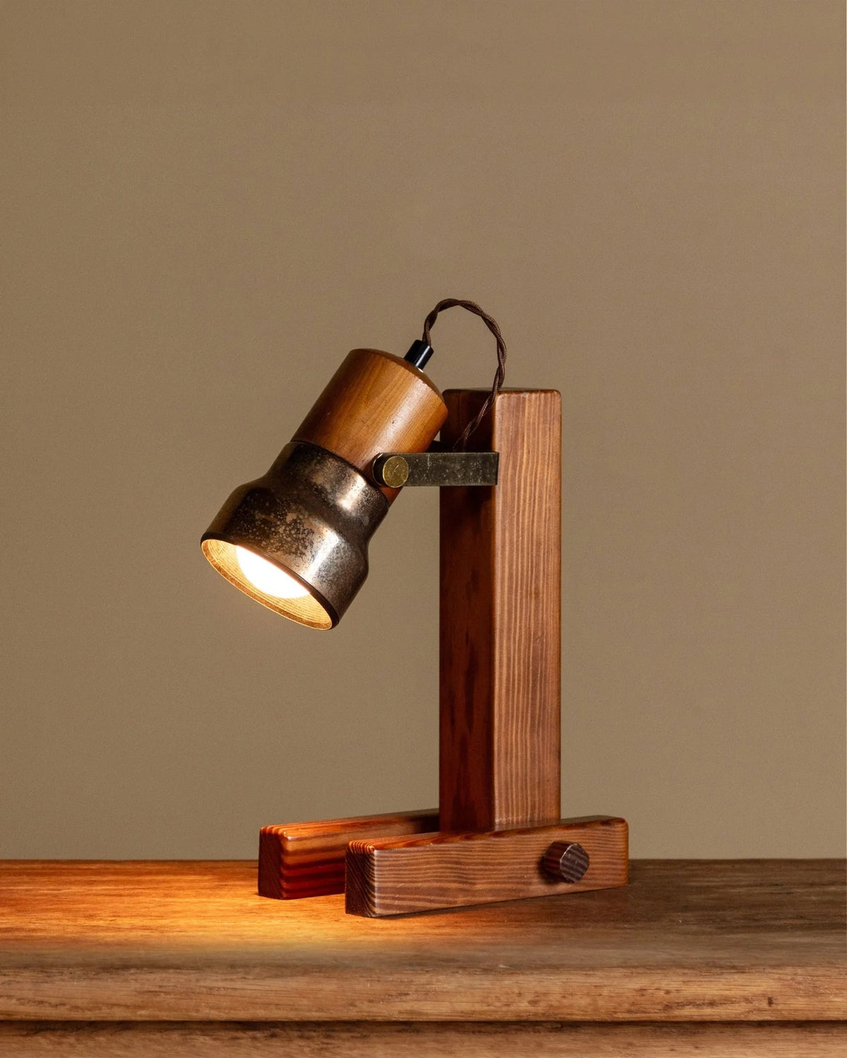 A rustic wooden table lamp with a warm metal shade and a dark wood stand, illuminated on a wooden surface against a solid beige background.