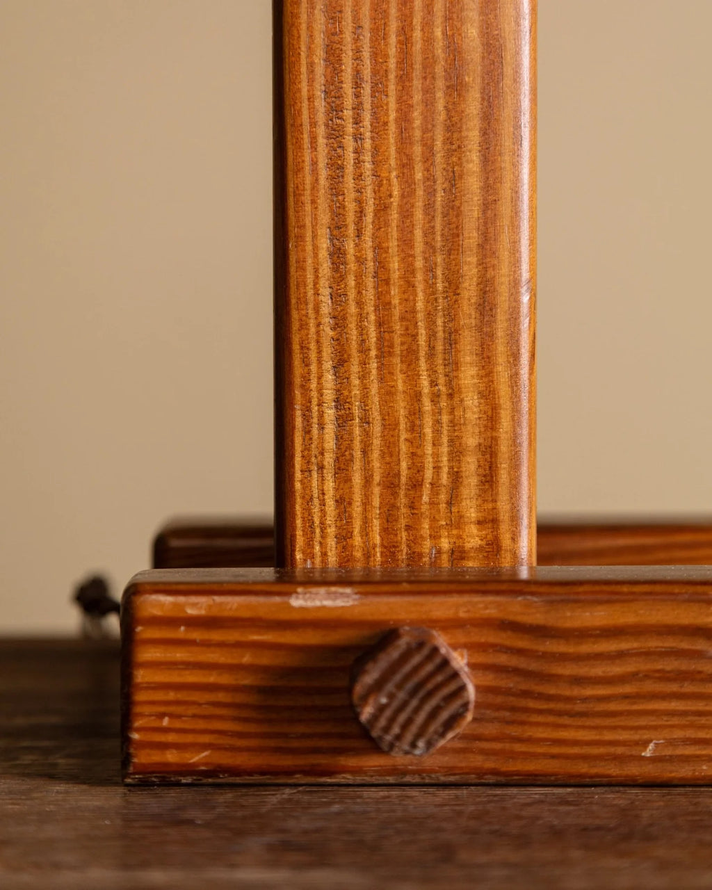 Close-up of a wooden furniture leg joint showing a polished medium-brown wood grain surface with a round turned wood knob, set against a neutral beige background.
