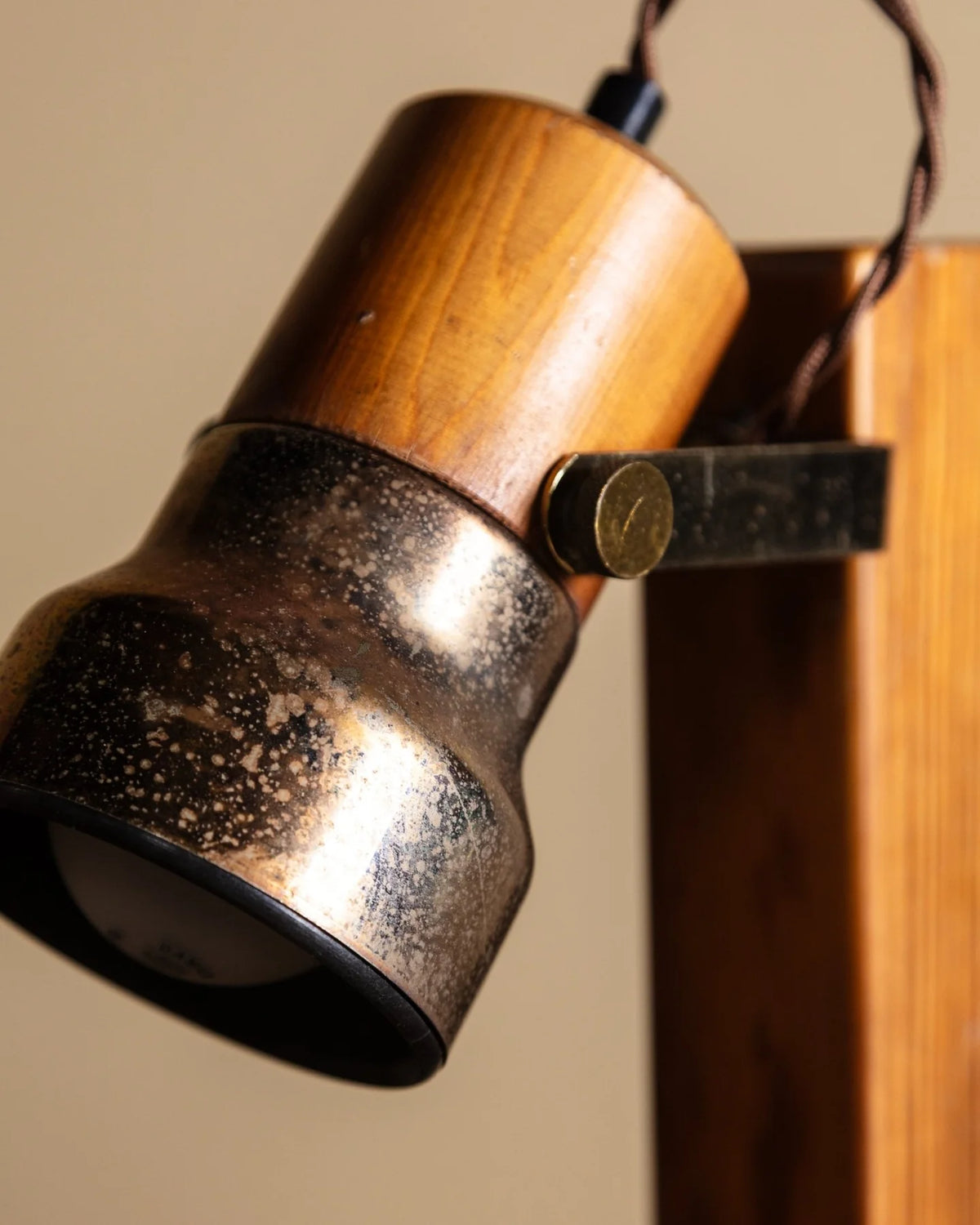 Close-up of a vintage-style industrial wall lamp with a brushed bronze metal shade, a natural wood cylinder, and a black metal bracket, shown against a warm beige background.