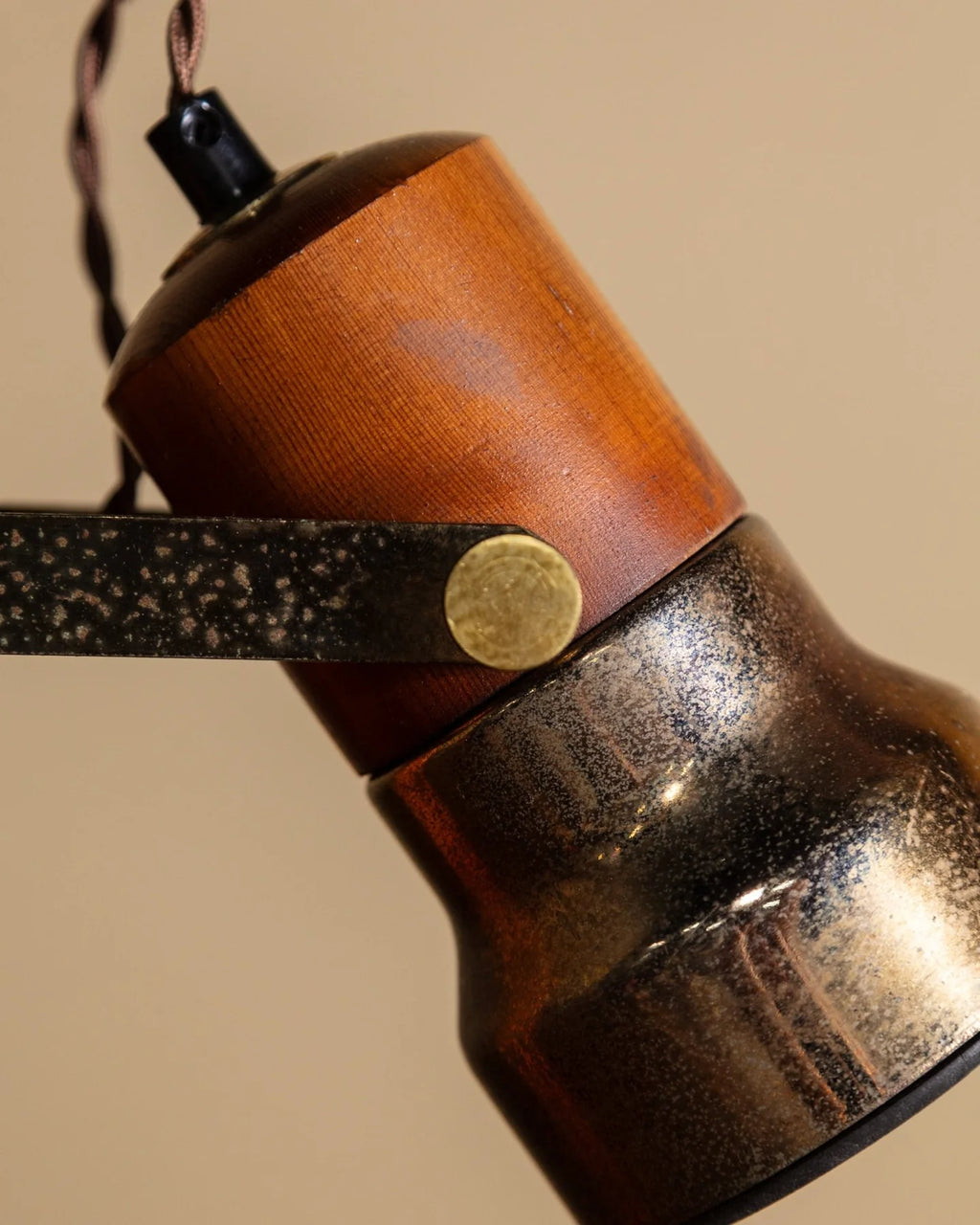 Close-up of a vintage-style industrial lamp shade featuring a warm brown wood top, a dark metallic bronze lower section, and a black metal support arm with a brass-colored rivet, set against a solid beige background.