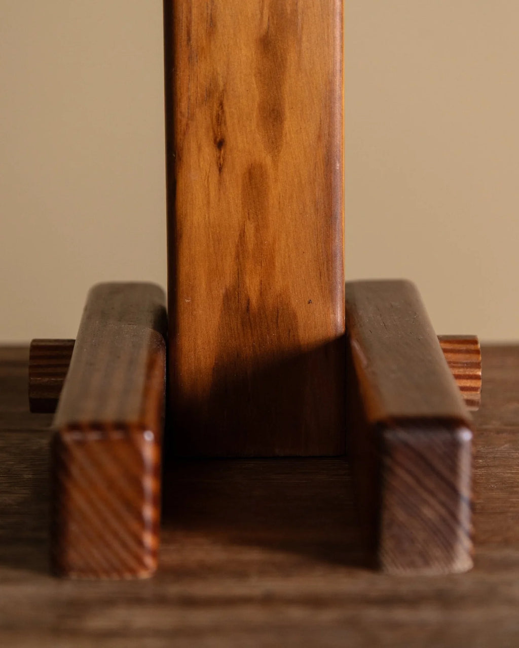 A close-up of a wooden display stand or easel with a vertical center panel and two horizontal support bars, shown on a wooden surface against a plain beige background.