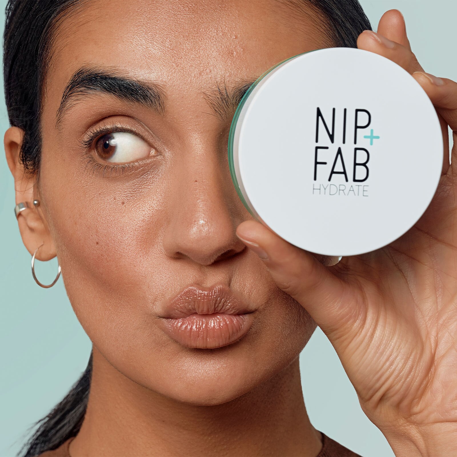 A close-up of a woman holding a round white NIP+FAB HYDRATE cosmetic jar against her eye, with a pale blue background.