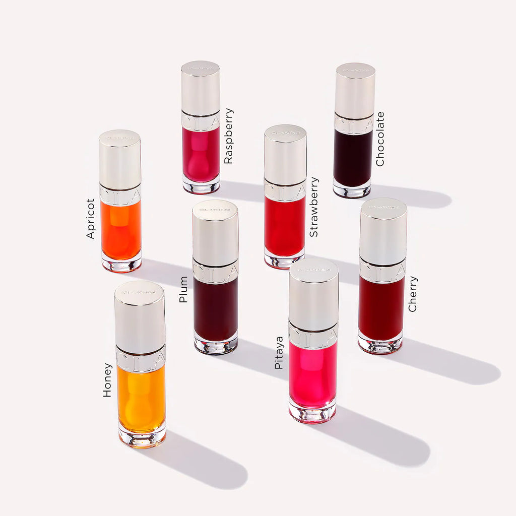 Nine lip gloss tubes arranged on a light beige background, each labeled with a flavor name: Apricot, Raspberry, Chocolate, Honey, Plum, Strawberry, Pilsa, and Cherry. The tubes have silver caps and clear glass bodies showing different gloss colors from bright pink and red to orange, deep burgundy, and dark brown.