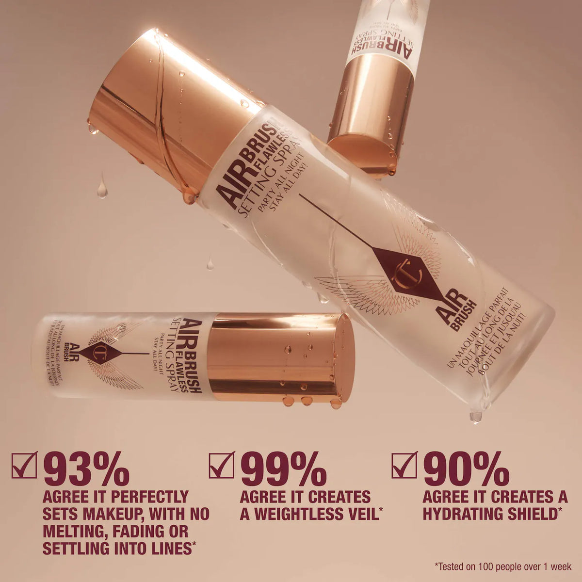 Three Airbrush Setting Sprays from Beauty Creations are arranged diagonally on a warm beige background with water droplets. The sprays are frosted white with metallic rose gold caps and feature a black and white label with the brand logo.