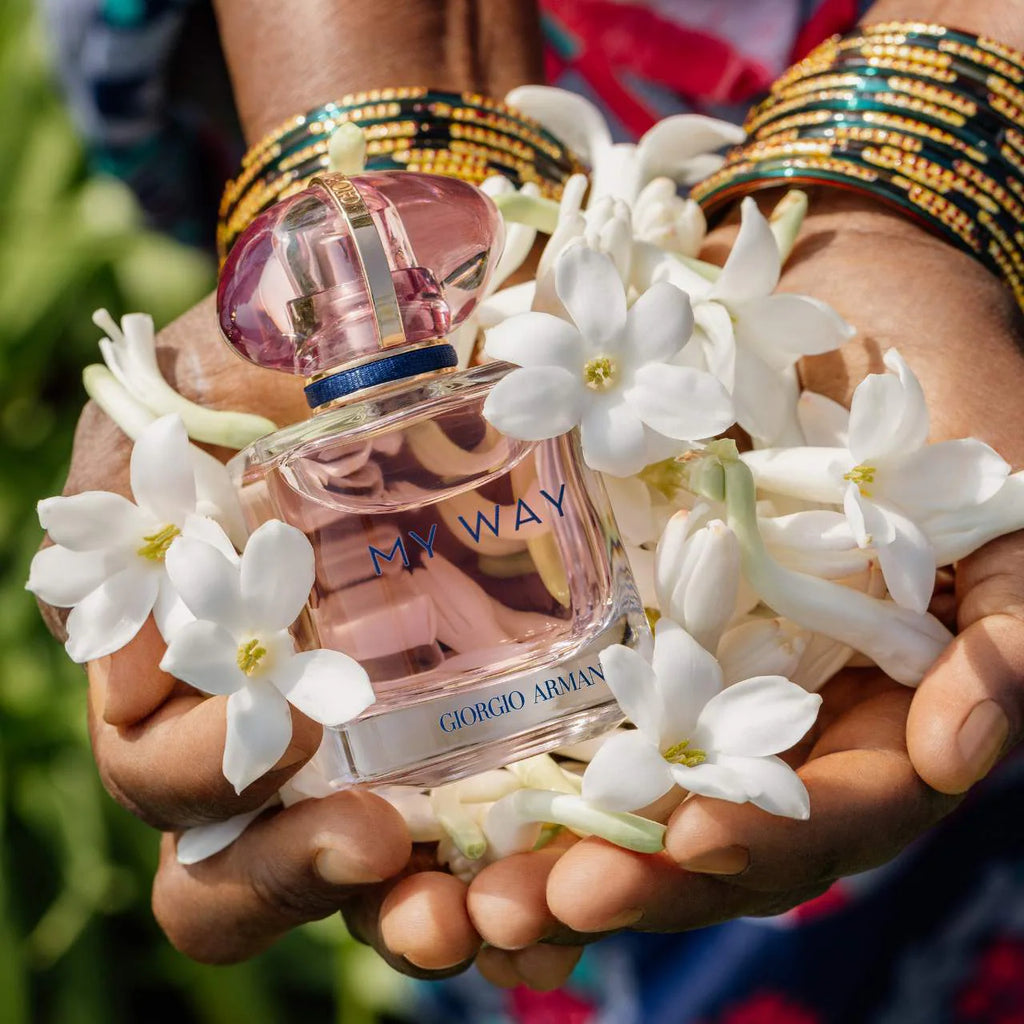A clear glass perfume bottle labeled 'MY WAY' and 'GIORGIO ARMANI' held in two hands surrounded by white flowers. The person wears multiple gold and green beaded bangles, with a blurred green and blue outdoor background.