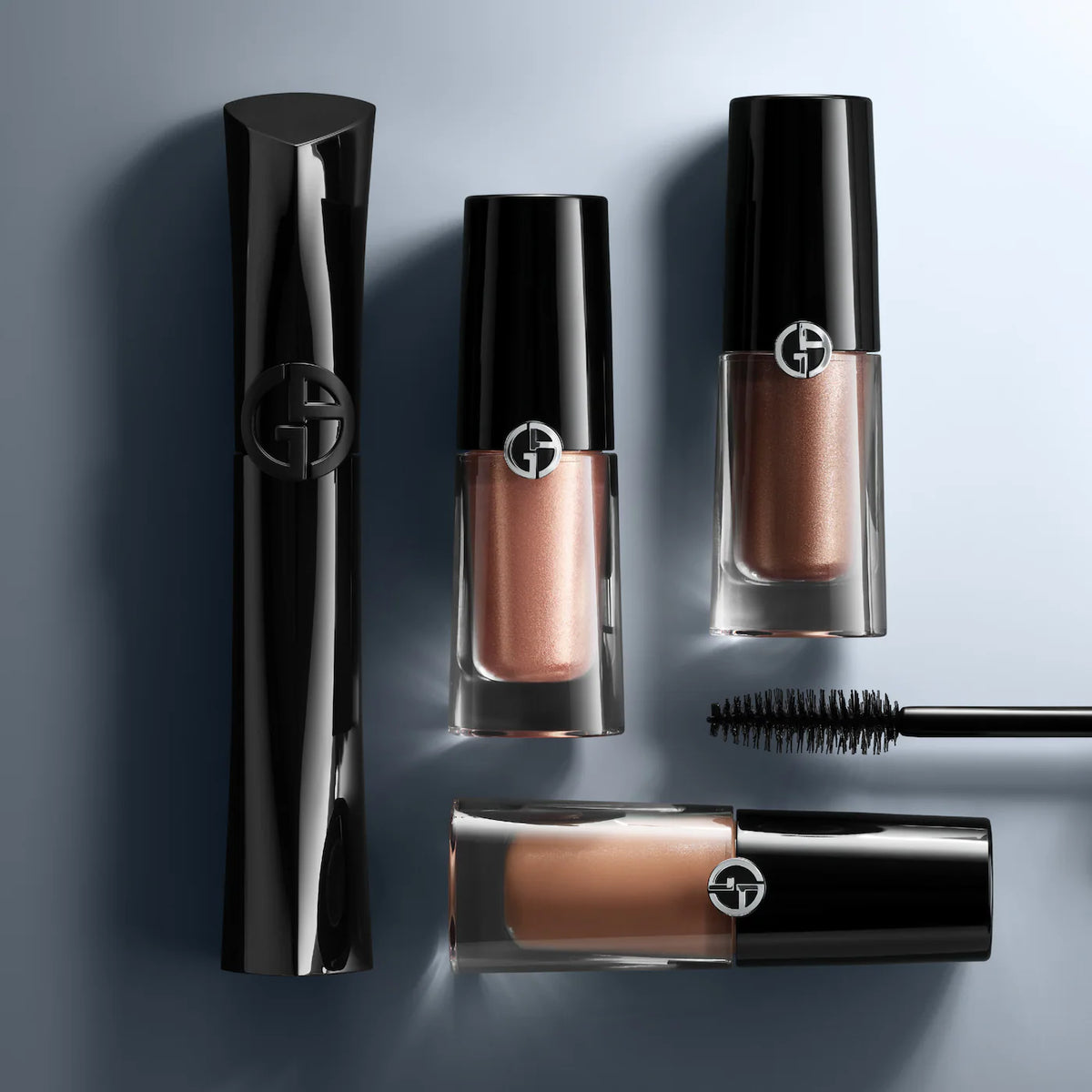Four Giorgio Armani cosmetic tubes and a mascara wand arranged on a cool blue-gray background. The set includes a tall black case, two black-and-clear lip products showing rose-gold and peachy nude shades, and a black mascara wand.