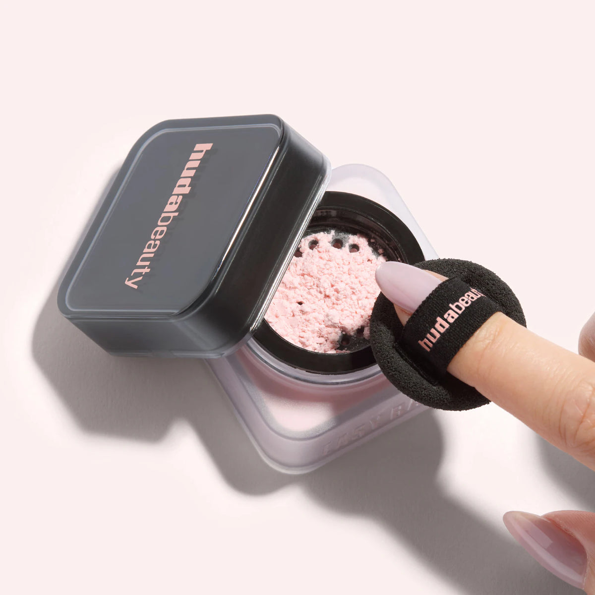 Huda Beauty compact powder with a pink powder and black sponge applicator on a light gray background.