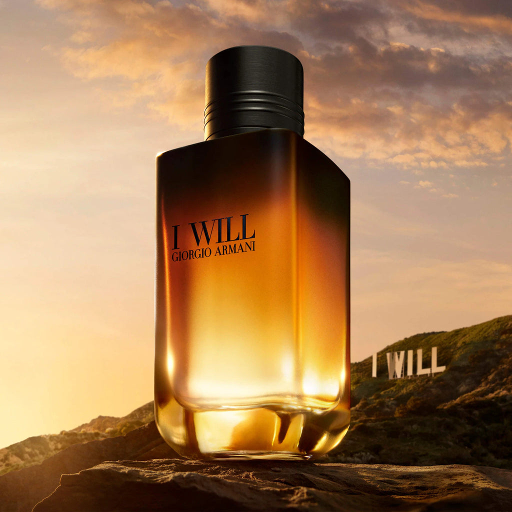 Giorgio Armani 'I WILL' perfume bottle, a faceted amber glass container with a black cap, set against a dramatic sunset sky and the Hollywood hills with the 'I WILL' sign.
