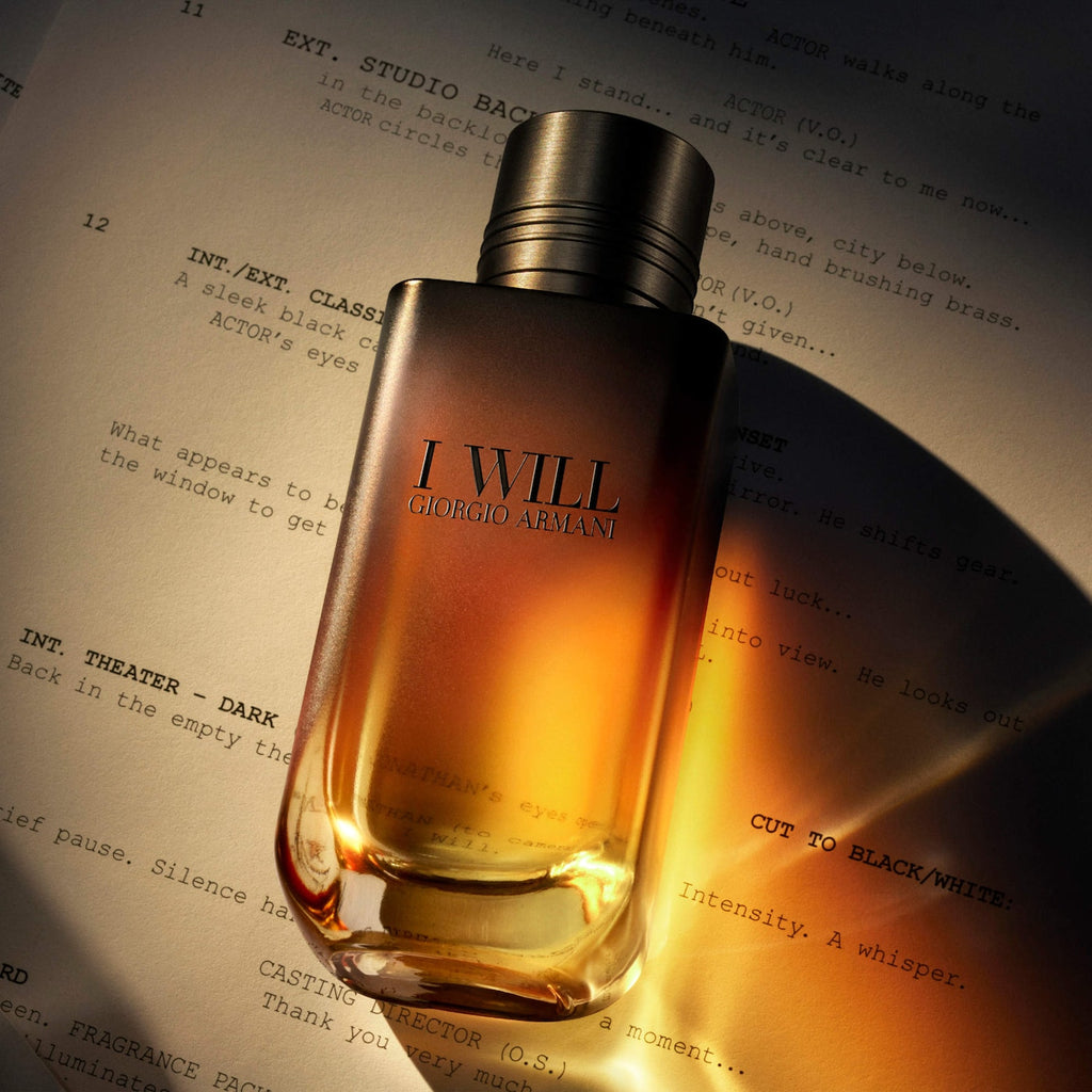 Giorgio Armani 'I Will' fragrance bottle with amber liquid and a metallic silver cap, resting on a printed script under dramatic warm lighting.