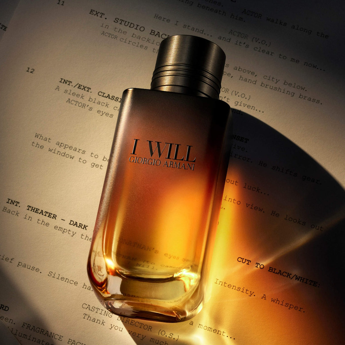 Giorgio Armani 'I Will' fragrance bottle with amber liquid and a metallic silver cap, resting on a printed script under dramatic warm lighting.