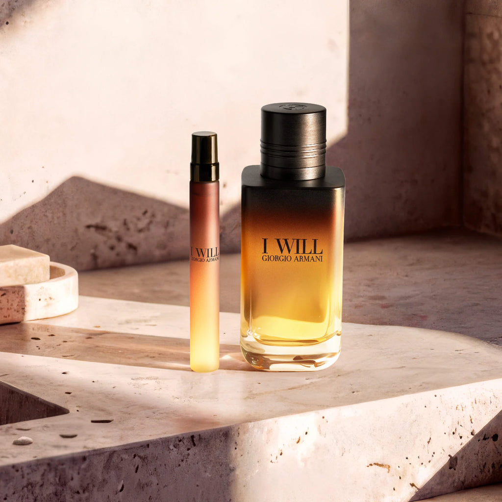 A bottle of Giorgio Armani 'I Will' fragrance and a matching travel spray vial, both featuring a warm amber-to-cream gradient, displayed on a textured stone ledge with dramatic warm lighting.
